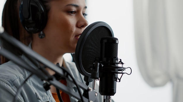 Young woman talking to audience while using professional microphone. Internet live radio talk show host presenting last news of day while sitting in studio with modern headphones on.