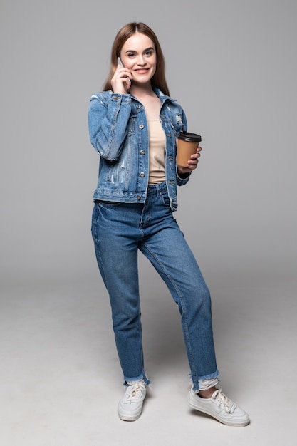 Young woman talking by mobile phone drinking coffee posing isolated over grey wall