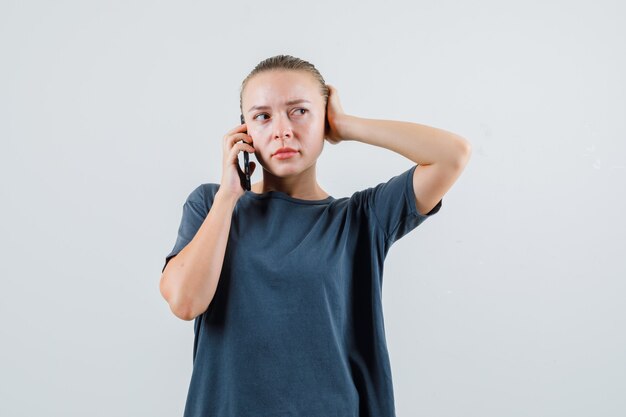 Молодая женщина разговаривает по мобильному телефону в серой футболке и выглядит задумчивой
