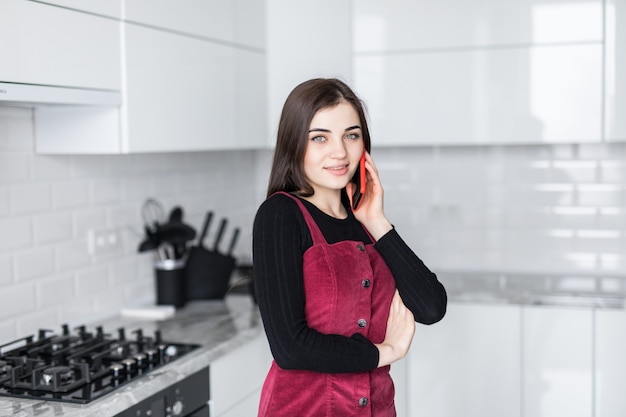 Молодая женщина разговаривает по мобильному телефону при использовании ноутбука на кухне у себя дома