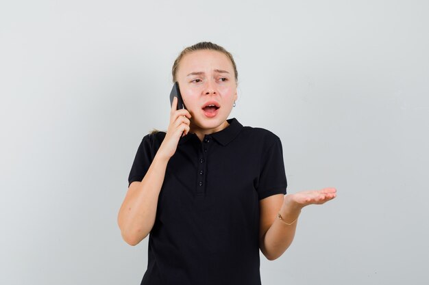 Молодая женщина разговаривает по телефону и поднимает одну руку в черной футболке и выглядит разочарованной