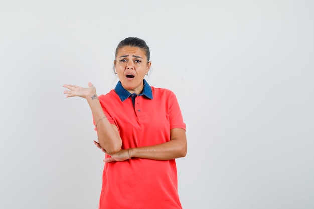 Young woman talking and stretching palm in red t-shirt and looking curious , front view.