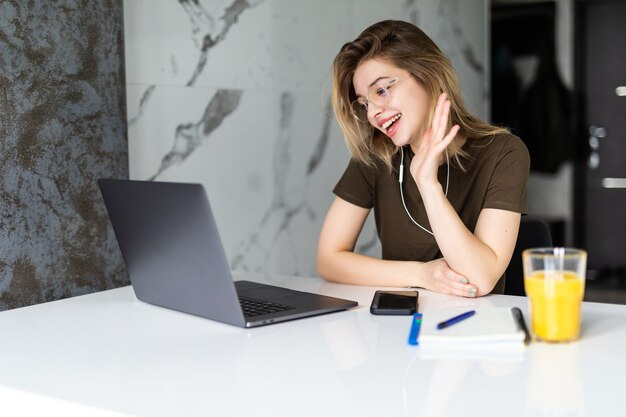 Young woman talking on video call and waving hand while sitting at table in the kitchen