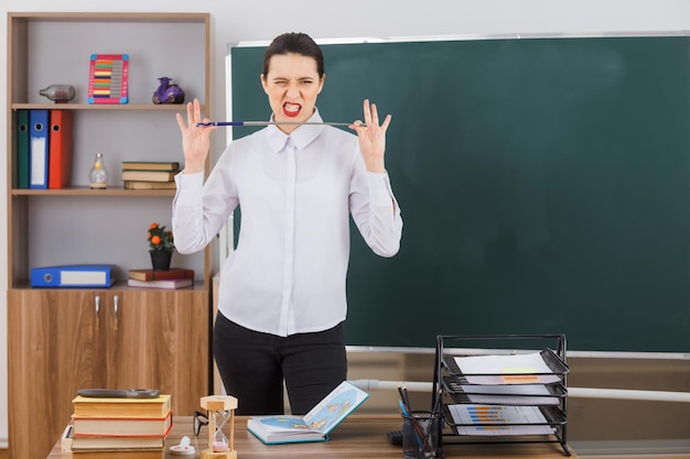 無料写真 ポインターを持っている若い女性教師は、教室の黒板の前にある学校の机に立って自信を持って怒っているように見えるレッスンを説明しようとしています