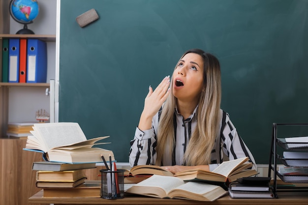 young woman teacher sitting at school desk in front of blackboard in classroom among books on her desk looking tired and overworked yawning covering mouth with hand