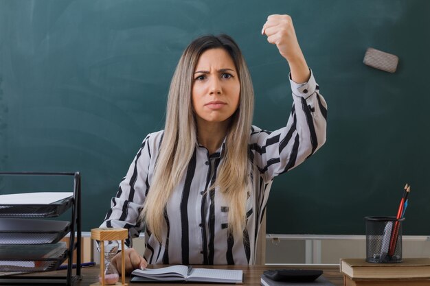 教室の黒板の前の学校の机に座っている若い女教師は、怒って怒って握りこぶしを上げる学生の宿題をチェックします