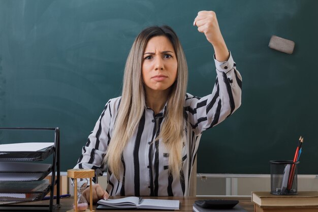 young woman teacher sitting at school desk in front of blackboard in classroom checking homework of students angry and mad raising clenched fist