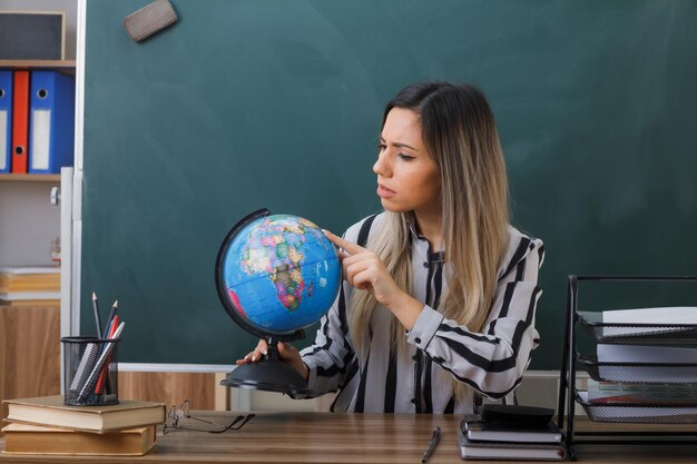 young woman teacher sitting at school desk in front of blackboard in classroom explaining lesson holding globe looking at it intrigued