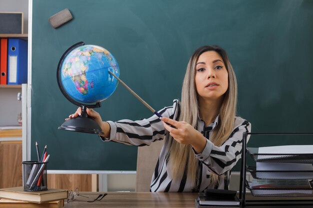 young woman teacher sitting at school desk in front of blackboard in classroom explaining lesson holding globe and pointer looking confident