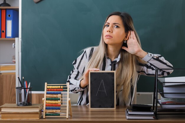 young woman teacher sitting at school desk in front of blackboard in classroom explaining lesson holding small chalkboard holding hand near ear trying to hear something