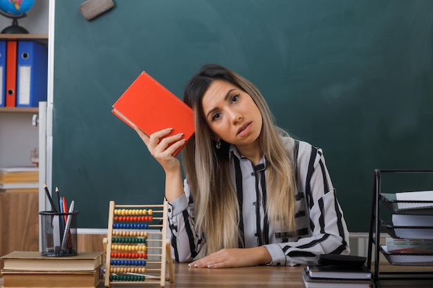 young woman teacher sitting at school desk in front of blackboard in classroom holding book looking puzzled