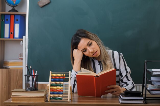 young woman teacher sitting at school desk in front of blackboard in classroom reading book looking tired and bored