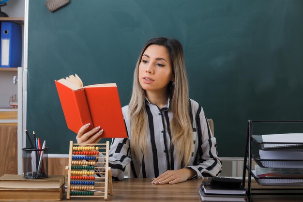 young woman teacher sitting at school desk in front of blackboard in classroom reading book preparing for lesson looking confident