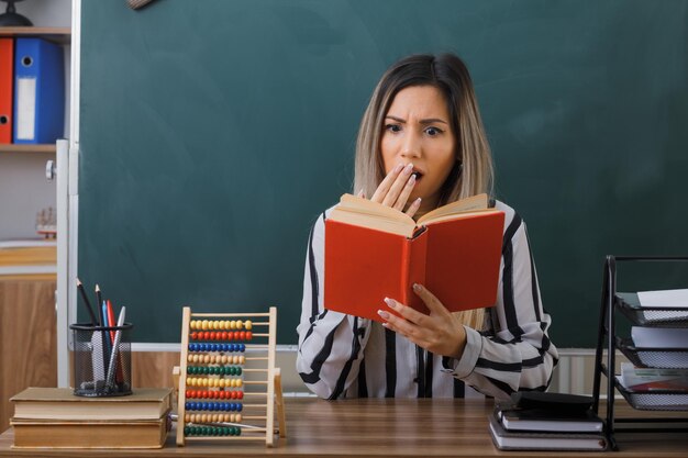 молодая женщина-учитель сидит за школьной партой перед доской в классе и читает книгу, готовясь к уроку, выглядит взволнованной и шокированной