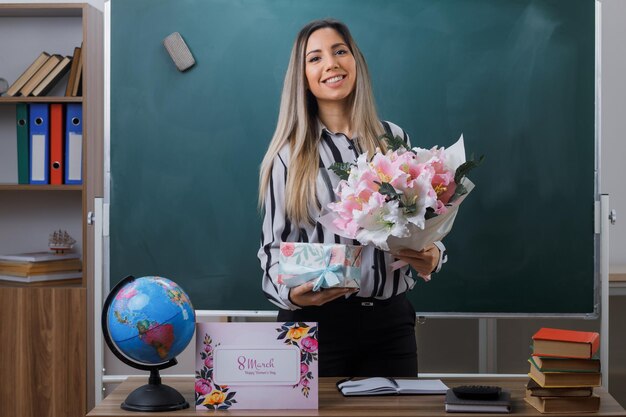 young woman teacher standing near blackboard in classroom holding flower bouquet and present box from students looking happy and pleased happy womens day concept