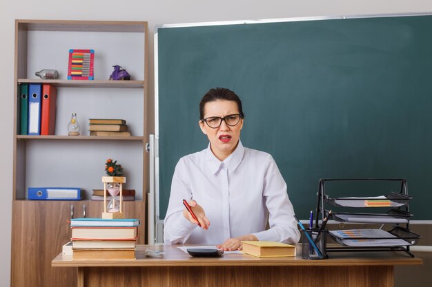 Young woman teacher wearing glasses checking homework of students being displeased sitting at school desk in front of blackboard in classroom