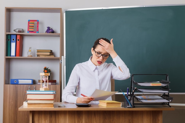 Young woman teacher wearing glasses checking homework of students looking confused with hand on her forehead sitting at school desk in front of blackboard in classroom