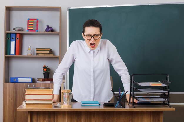 眼鏡をかけた若い女教師が教室の黒板の前の学校の机に座って怒って欲求不満の授業を説明する