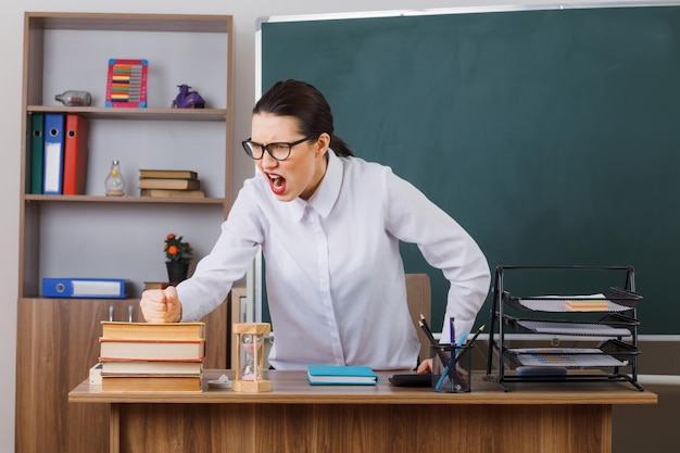 Insegnante di giovane donna con gli occhiali che spiega la lezione arrabbiata e frustrata che impazzisce seduto al banco della scuola davanti alla lavagna in classe