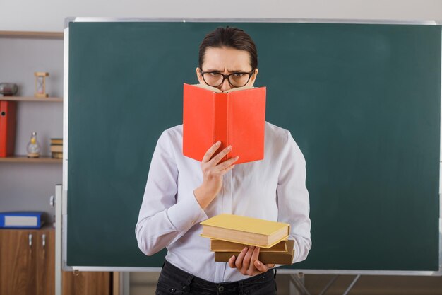 Young woman teacher wearing glasses explaining lesson holding stack of books frowning being displeased standing at school desk in front of blackboard in classroom