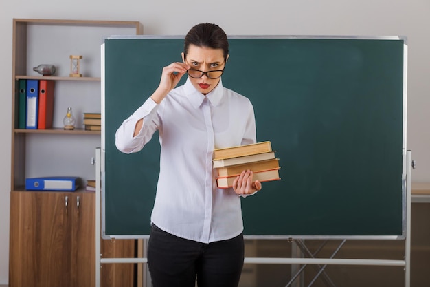 Young woman teacher wearing glasses explaining lesson holding stack of books frowning standing at school desk in front of blackboard in classroom