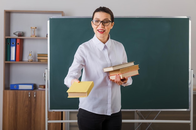 Young woman teacher wearing glasses explaining lesson holding stack of books smiling confident standing at school desk in front of blackboard in classroom