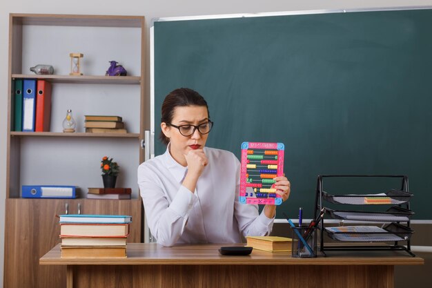 そろばんを持った眼鏡をかけた若い女教師が、教室の黒板の前にある学校の机に座って困惑した考えでレッスンを説明