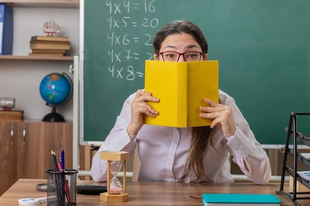 Young woman teacher wearing glasses holding book covering face with it preparing for lesson sitting at school desk in front of blackboard in classroom