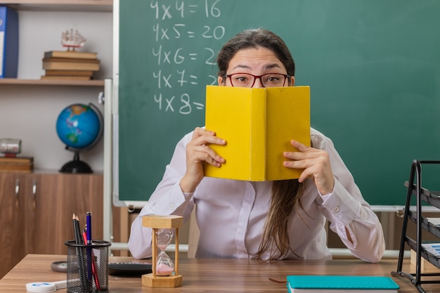 Молодая женщина-учитель в очках держит книгу, закрывающую лицо, готовится к уроку, сидя за школьной партой перед доской в классе
