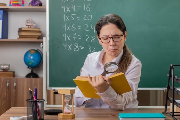 Young woman teacher wearing glasses holding book looking at pages through magnifying glass with confident expression sitting at school desk in front of blackboard in classroom