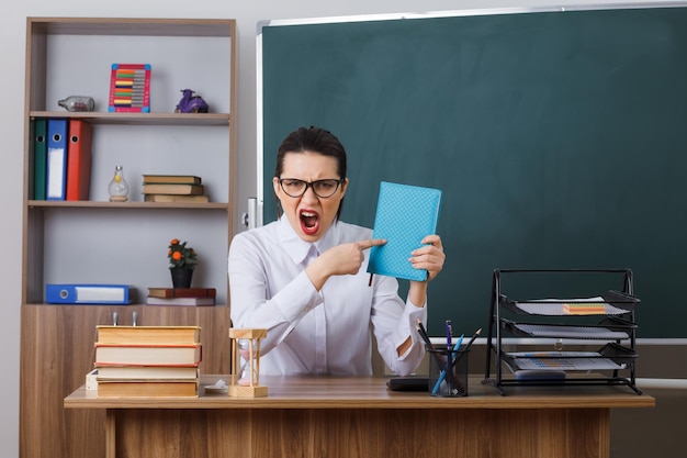 Insegnante di giovane donna con gli occhiali che tiene il libro che punta con il dito indice verso di esso gridando arrabbiato e frustrato seduto al banco di scuola davanti alla lavagna in classe