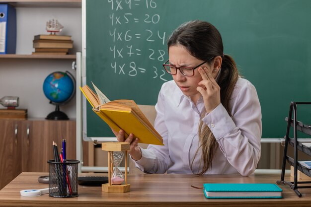 Young woman teacher wearing glasses holding book preparing for lesson reading being confused