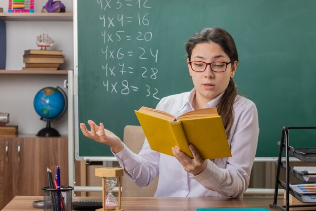 교실에서 칠판 앞에 학교 책상에 앉아 수업 독서를 준비하는 책을 들고 안경을 착용하는 젊은 여자 교사