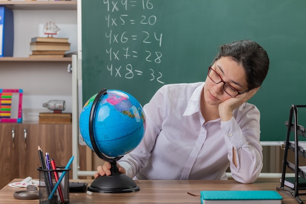 Young woman teacher wearing glasses holding globe looking at it being tired