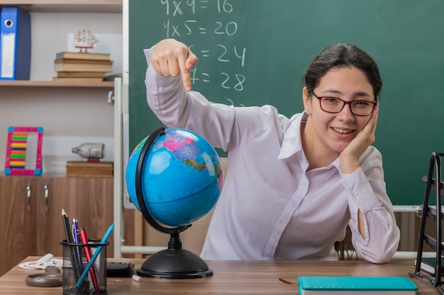 Young woman teacher wearing glasses holding globe pointing with index finger at it smiling cheerfully explaining lesson sitting at school desk in front of blackboard in classroom