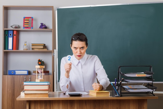 Insegnante di giovane donna con gli occhiali che tiene la targa due spiegando la lezione con la faccia accigliata che stringe il pugno seduto al banco della scuola davanti alla lavagna in aula