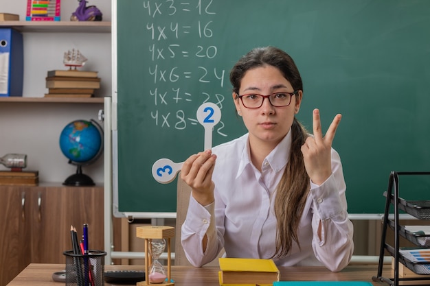 Young woman teacher wearing glasses holding number plates explaining lesson showing number two with fingers looking confident sitting at school desk in front of blackboard in classroom