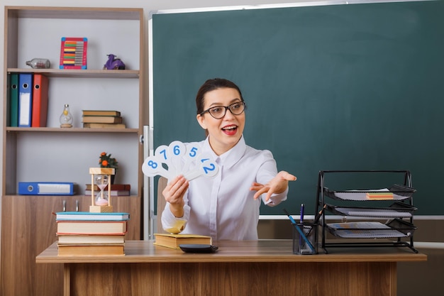 수업을 설명하는 번호판을 들고 안경을 쓴 젊은 여성 교사는 교실 칠판 앞에 자신감 있게 앉아 웃고 있다