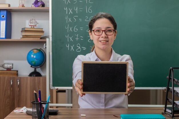 Young woman teacher wearing glasses holding small blackboard explaining lesson smiling confident sitting at school desk in front of blackboard in classroom