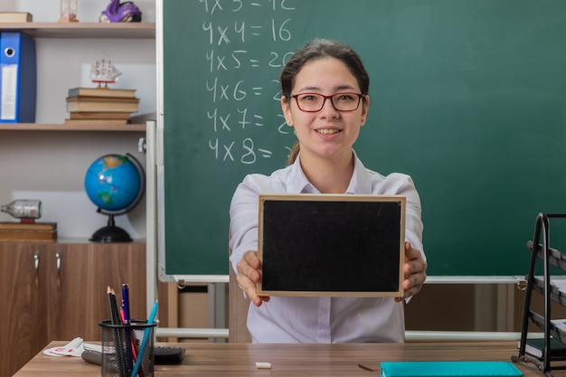 小さな黒板を持って眼鏡をかけている若い女性教師が教室の黒板の前にある学校の机に座って自信を持って笑顔でレッスンを説明します