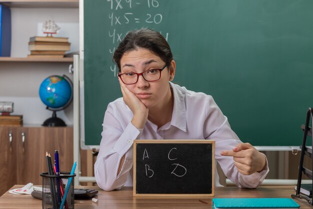 교실에서 칠판 앞에 학교 책상에 앉아 불쾌한 찾고 수업을 설명하는 그것에 검지 손가락으로 가리키는 작은 칠판을 들고 안경을 착용하는 젊은 여자 교사