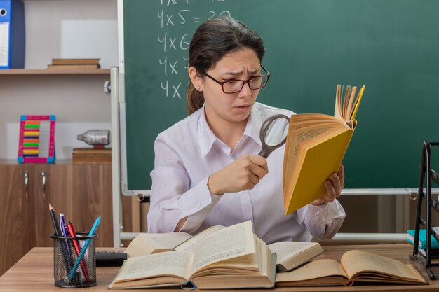 Young woman teacher wearing glasses looking at book through magnifying glass being confused and displeased sitting at school desk in front of blackboard in classroom