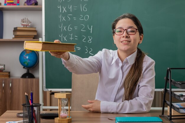 Young woman teacher wearing glasses offering book smiling confident sitting at school desk in front of blackboard in classroom