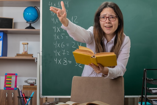молодая женщина-учитель в очках читает книгу, объясняя урок, протягивая руку, смущенная и недовольная, стоя за школьной партой перед доской в классе