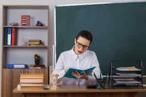 무료 사진 교실에서 칠판 앞에 앉아 수업을 준비하는 진지한 얼굴로 책을 읽는 안경을 쓴 젊은 여성 교사