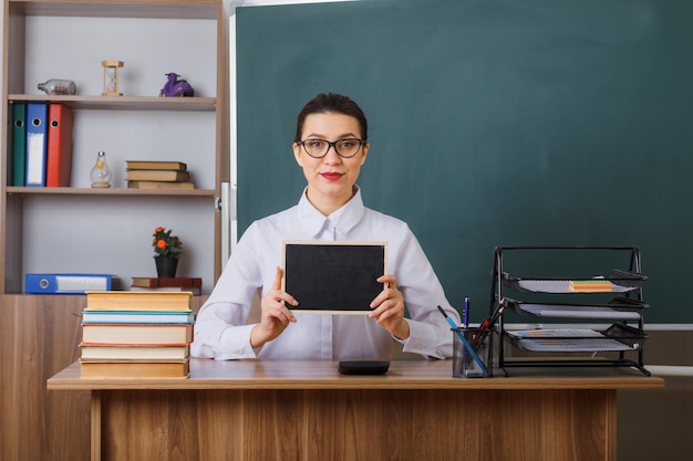 Foto gratuita insegnante di giovane donna con gli occhiali che mostrano una piccola lavagna sorridente sicura seduta al banco di scuola davanti alla lavagna in classe