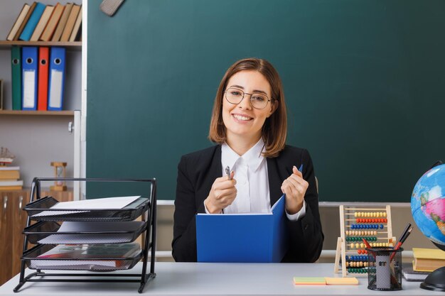 Young woman teacher wearing glasses sitting at school desk in front of blackboard in classroom checking class register looking at camera smiling cheerfully