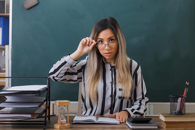 young woman teacher wearing glasses sitting at school desk in front of blackboard in classroom checking homework of students looking at camera confused