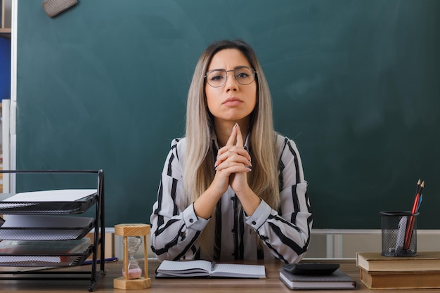 young woman teacher wearing glasses sitting at school desk in front of blackboard in classroom checking homework of students looking at camera with serious face holding hands together