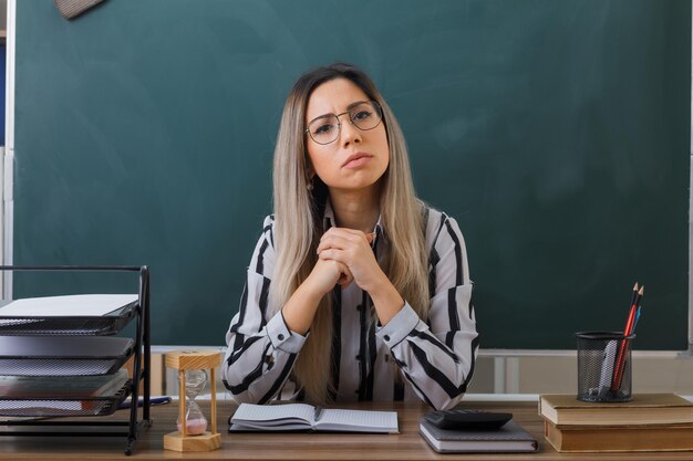 young woman teacher wearing glasses sitting at school desk in front of blackboard in classroom checking homework of students looking at camera with serious face holding hands together